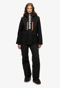 Czarna kurtka i spodnie narciarskie z pomarańczowymi akcentami; posiada logo, zamki błyskawiczne, kieszenie oraz materiał wodoodporny; zaprojektowane do zimnej pogody.