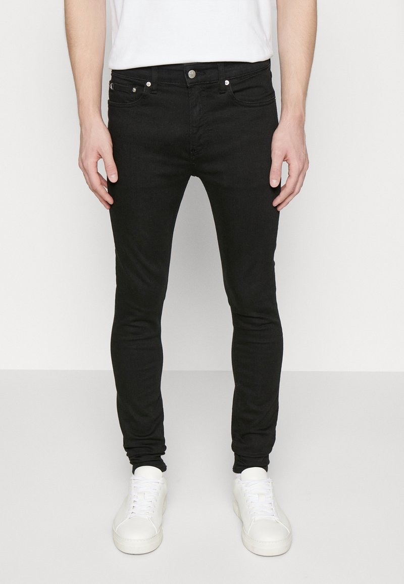 Hombre con jeans negros ajustados y zapatillas blancas de pie contra un fondo claro y liso, con las manos relajadas a los lados.