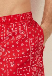 Žluté plavky s potiskem červených bandan, které mají bílé paisley vzory, elastický pas a boční kapsy. Hladký textilní povrch.