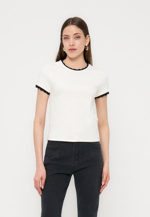 Donna con indosso una maglietta bianca a maniche corte con bordi smerlati neri e pantaloni neri a vita alta, in piedi davanti a uno sfondo chiaro e uniforme.