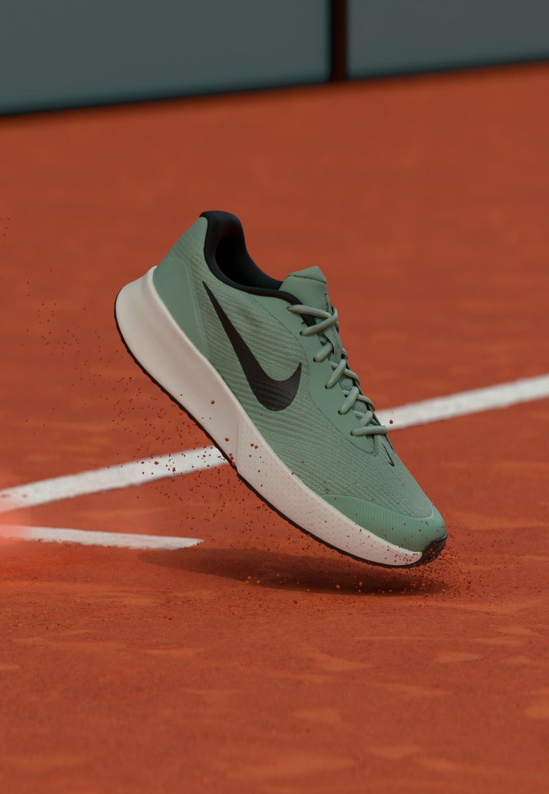 Zapatilla de correr Nike verde claro con el logo negro, suspendida en el aire sobre una pista de tenis de arcilla roja con líneas blancas.