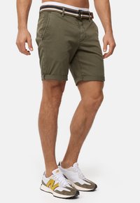 Shorts en coton vert olive avec des revers, ornés d'une ceinture à rayures multicolores et d'un design de poche classique. Portés avec des baskets blanches et grises.