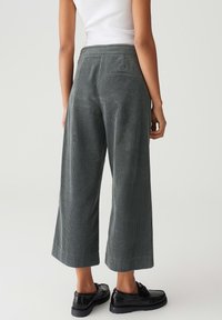 Personne portant un pantalon cropped large en velours côtelé gris taille haute, associé à des mocassins noirs et un haut sans manches blanc, debout.