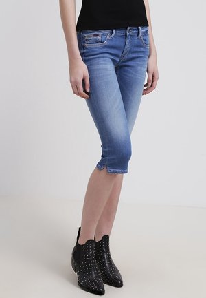 Smaltsiddende blå denim capri bukser båret med sorte nitte-støvler og en sort top, vist fra taljen til fødderne mod en ensfarvet baggrund.