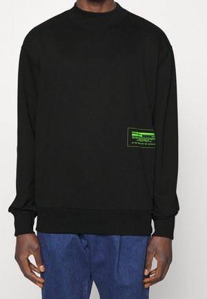Sweatshirt noir en tissu doux, avec un col rond, des poignets côtelés et une étiquette graphique verte sur le côté gauche.