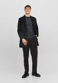 Manteau noir sur un pull gris foncé, associé à un pantalon et des chaussures noires. Le modèle se tient avec les mains dans les poches devant un fond clair.