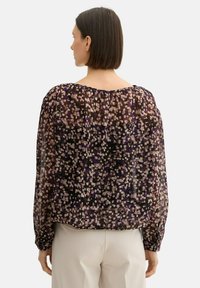 Durchsichtige Bluse mit langen Ärmeln, die einen schwarzen Grundton und ein farbenfrohes, abstraktes Muster aus lila, beige und weißen Punkten aufweist.