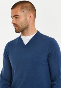 Blauer V-Ausschnitt-Pullover aus strukturiertem Strickstoff mit langen Ärmeln und tailliertem Design, getragen über einem weißen T-Shirt.