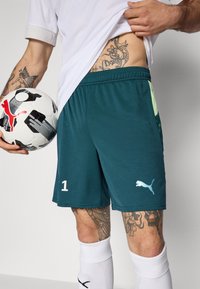 Turkooise sportshorts met een elastische tailleband, een wit nummer "1" en het Puma-logo. Een gestructureerde afwerking met lichte groene accenten is zichtbaar.
