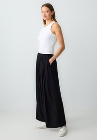 Pantalons larges plissés noirs avec poches latérales, associés à un débardeur blanc ajusté. Le modèle se tient devant un fond gris clair.