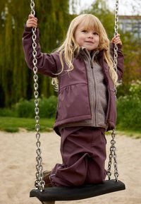 Enfant sur une balançoire portant une veste violette avec une doublure en polaire et un pantalon violet assorti. La balançoire a un siège noir et un support en chaîne en métal.