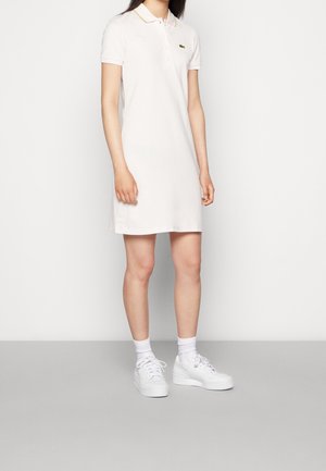 Kobieta ubrana w biały, krótki, koszulowy strój polo z małym zielonym logo na piersi, białe skarpetki i białe trampki, stojąca na szarej podłodze.