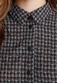 Camicia a quadri marrone e blu con colletto a punta, bottoni neri e tessuto texture. Primo piano dei dettagli anteriori.