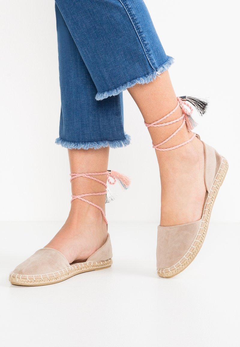 Ballerines espadrilles beiges avec des lacets roses à nouer autour de la cheville et des pompons, portées avec un jean bleu coupé à l'ourlet effiloché.