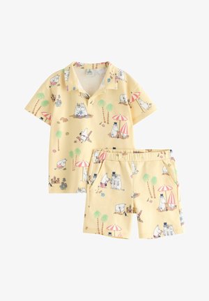Lasten keltainen lyhythihainen paita ja shortsit, joissa on sarjakuvatiivisteisiä virtahepohahmoja, palmuja, aurinkovarjoja ja hiekkalinnakuvioita rannalla.