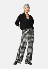Veste bomber en dentelle noire sur un haut noir à col en V, associée à un pantalon large à motifs géométriques noir et blanc et des sandales noires.