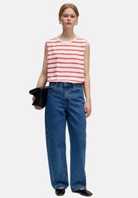 Weißes ärmelloses Oberteil mit roten Streifen, kombiniert mit lockeren blauen Jeans. Model hält eine schwarze Clutch. Glatte Texturen und lässiges Design.