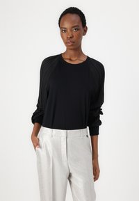 Chemise noire avec des manches bouffantes texturées, accompagnée de pantalons argentés brillants. La chemise possède un col rond et une coupe décontractée.