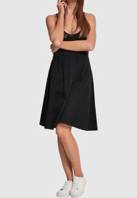 Svart ärmlös klänning med utsvängd kjol, tillverkad av mjukt tyg. Har en enkel design och slutar ovanför knäet, kombinerad med vita sneakers.