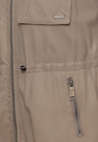 Beige leren jack met een frontale rits, een horizontale zak en een decoratieve logo plaat; beschikt over een gladde textuur en gedetailleerde stiksels.