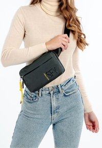 Texturerad svart axelväska med en gul accent, dragkedjestängning och två snedställda framfickor, bärs mot en beige topp och denimjeans.