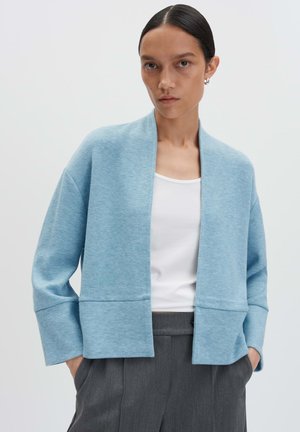 Cardigan aperto blu chiaro in morbido tessuto con maniche lunghe, vestibilità rilassata e dettagli di cuciture orizzontali, abbinato a una maglia bianca.