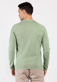 Uomo di spalle che indossa una camicia a maniche lunghe di un verde chiaro testurizzato e pantaloni beige su uno sfondo bianco uniforme.