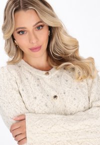 Jasno beżowy sweter zapinany na guziki z dzianiny, ozdobiony diamentowymi wzorami, srebrnymi dodatkami i złotymi guzikami, z długimi rękawami i ściągaczami.