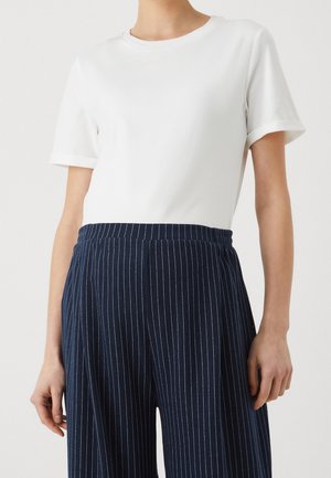 Vrouw draagt een effen wit kortermig shirt, ingestopt in marineblauwe broek met wijde pijpen en krijtstrepen, tegen een neutrale achtergrond.