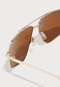 Gafas de sol tipo aviador con lentes marrones, montura de metal dorado, detalles acanalados en el ceño y almohadillas nasales transparentes.