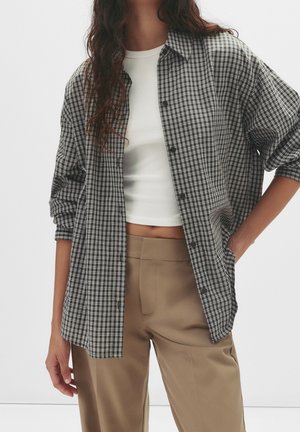 Femme portant une chemise à carreaux noire et blanche boutonnée sur un crop top blanc et un pantalon beige taille haute, main dans la poche, sur fond blanc.