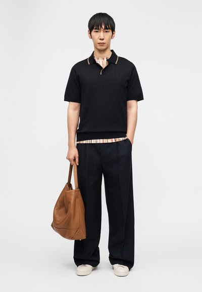 Νεαρός άνδρας που στέκεται, φορώντας μαύρο κοντομάνικο πουκάμισο πάνω από ριγέ τοπ, μαύρο παντελόνι φαρδιάς γραμμής, λευκά αθλητικά παπούτσια και κρατώντας καφέ δερμάτινη τσάντα ώμου.