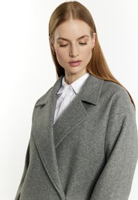 Grijze wollen jas met een notch-kraag, oversized pasvorm en zichtbare textuur. Daaronder is een witte blouse met kraag gedeeltelijk zichtbaar.