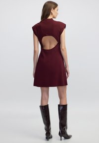Krátké burgundské šaty s krátkými rukávy, se spojeným velkým výřezem na zádech. Doplněné o vysoké, lesklé černé boty. Hladká struktura tkaniny.