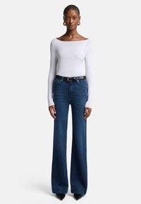 Top bianco a maniche lunghe con scollatura ampia abbinato a jeans svasati blu scuro, completato con una cintura nera e tacchi a punta neri.