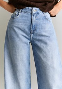 Persona che indossa jeans a gamba larga a vita alta di colore azzurro chiaro, con le mani infilate nelle tasche e un top marrone lavorato a maglia.