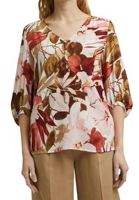 Blusa floreale con scollatura a V, caratterizzata da maniche ampie e gonfie. Il tessuto presenta un mix di motivi floreali rossi, rosa e marroni su uno sfondo chiaro.