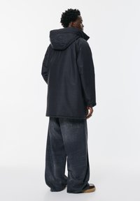 Bărbat îmbrăcat cu o jachetă neagră cu glugă și blugi gri închis, stând de-a latul pe un fundal alb simplu.