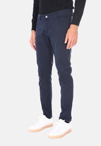 Uomo con pantaloni blu navy slim-fit e sneakers bianche, in piedi contro uno sfondo chiaro e uniforme.