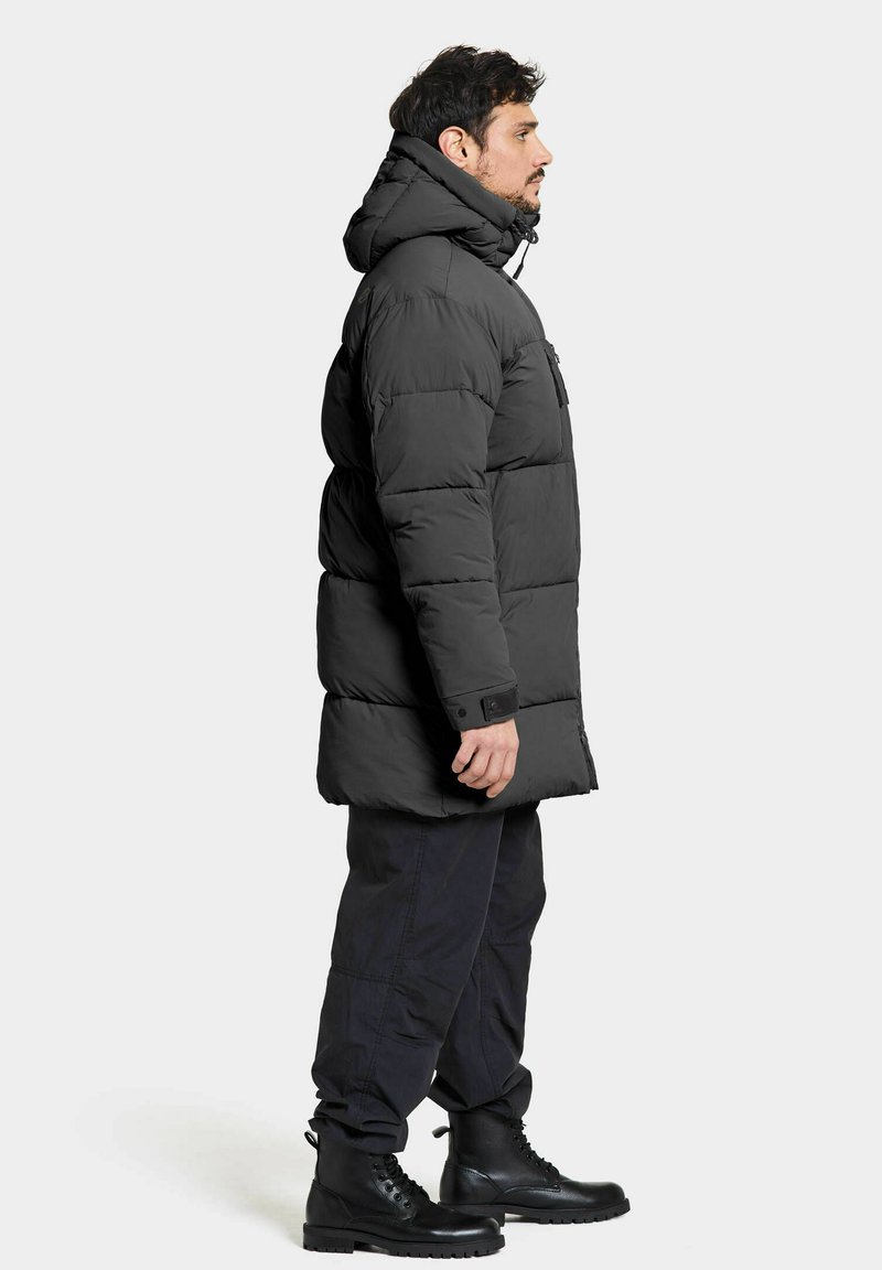 Schwarze Daunenjacke mit Kapuze, mit einem Reißverschluss vorne und einer Brusttasche, kombiniert mit dunklen Hosen und schwarzen Schnürstiefeln.