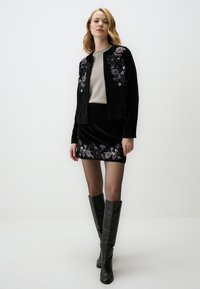 Completo giacca e gonna in velluto nero con ricami floreali in viola e bianco. Modella che indossa stivali neri fino al ginocchio e collant trasparenti.