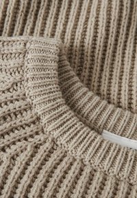 Maglione lavorato a maglia beige, vista ravvicinata. Texture a coste spesse, colletto rotondo, con un motivo uniforme su tutta la superficie e un piccolo etichetta visibile.