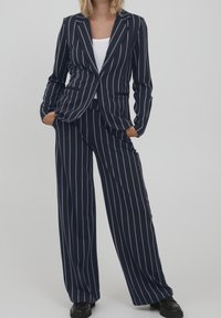 Femme debout les mains dans les poches, portant un blazer à rayures bleu marine et un pantalon large assorti, assortis à des chaussures noires.