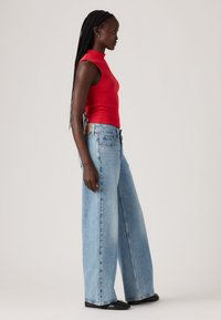 Rode mouwloze top met hoge hals, gecombineerd met lichtblauwe wijde jeans. Klassiek denim met een losse pasvorm en een hoge taille. Zwarte schoenen.