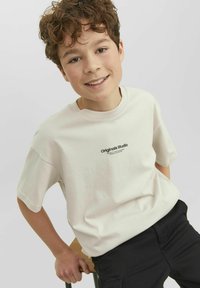 Ljusbeige bomulls t-shirt med korta ärmar har en svart logotyp med texten "Originals Studio" på bröstet och är kombinerad med svarta byxor.