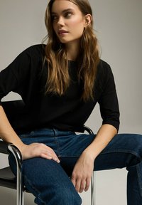 Czarny top z okrągłym dekoltem i rękawami o długości trzy czwarte, w połączeniu z niebieskimi dżinsami. Model siedzący na chromowanych krzesłach, przy delikatnym oświetleniu.