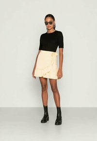 Mulher vestindo uma blusa preta de mangas curtas, saia drapeada creme com babado e laço, botas de tornozelo pretas e óculos escuros escuros, em pé contra um fundo simples.