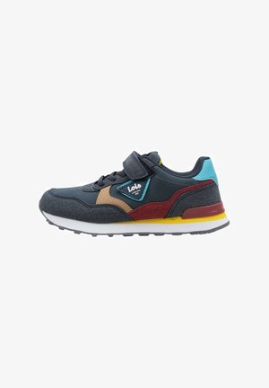 Zapatilla textil gris con una correa de Velcro, que presenta acentos en marrón, rojo y verde azulado, y una suela blanca con detalles en amarillo.