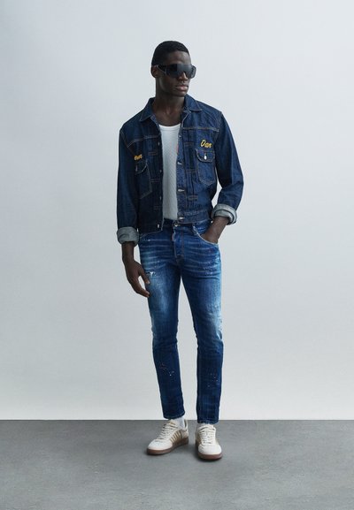 Uomo con giacca di jeans scura e jeans, canottiera bianca, sneakers bianche e occhiali da sole neri oversize, in piedi davanti a uno sfondo grigio uniforme.