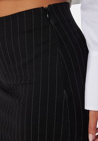 Jupe crayon noire avec de fines rayures blanches, dotée d'une fermeture éclair sur le côté et d'un design élégant et ajusté. Le matériau semble lisse et extensible.